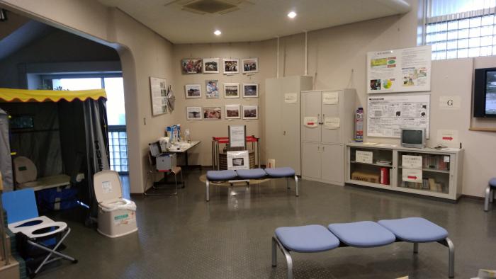 簡易トイレ、テント、3人掛けの椅子、パネル展示などが設置された室内の写真