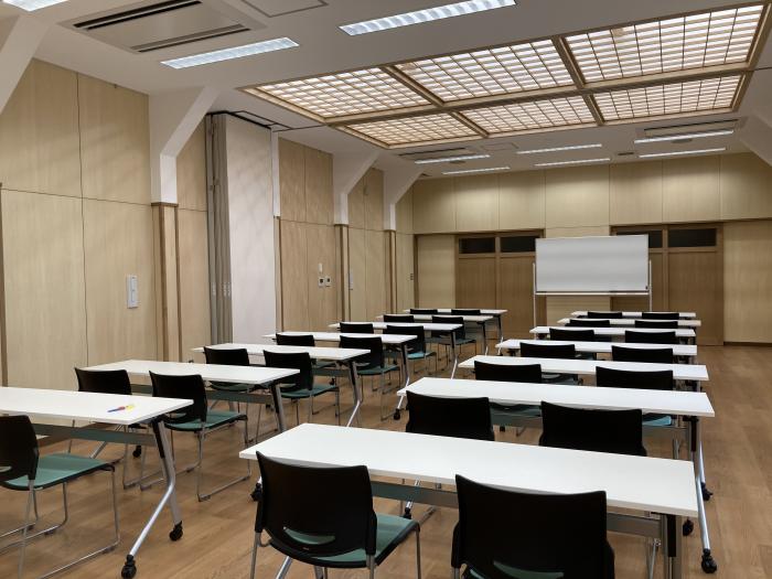 正面にホワイトボードが置かれ、白い長机がいくつも並べられた、格子状に仕切られている天井のライトが特徴的な研修室の写真