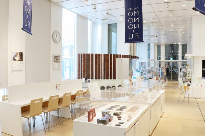 工芸品やクラフト品が展示されているいくつかの白いショーケースと5人が向かい合って座って休むことのできる細長い机が設置され、高い天井と細長い窓からの光で開放的な雰囲気のギャラリー「MONONO-FU」の写真