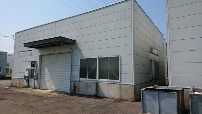 シャッターのある出入口を正面にパネル張りの白い壁で四面四角に囲うように建てられている工場棟の外観写真