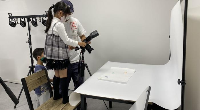 椅子の上に立った女児が男性に教わりながらカメラで写真を撮っている写真