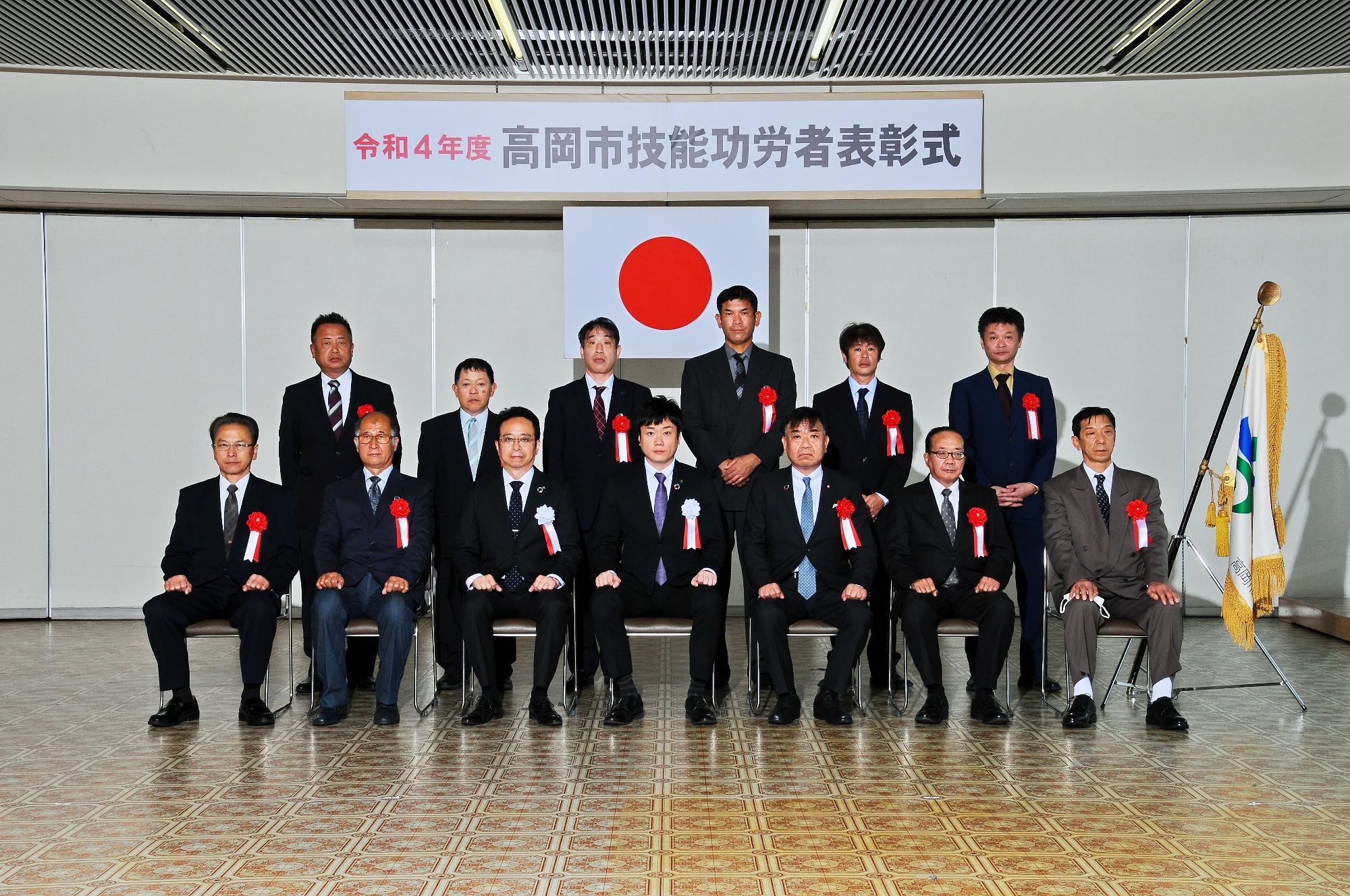 「令和4年度高岡市技能功労者表彰式」と書かれた横断幕と日本国旗の前に並ぶスーツ姿の人たちの集合写真