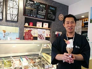 ジェラート店のショーケース横のカウンター前で大きなデザートを手に持った清都 勢憲さんの写真