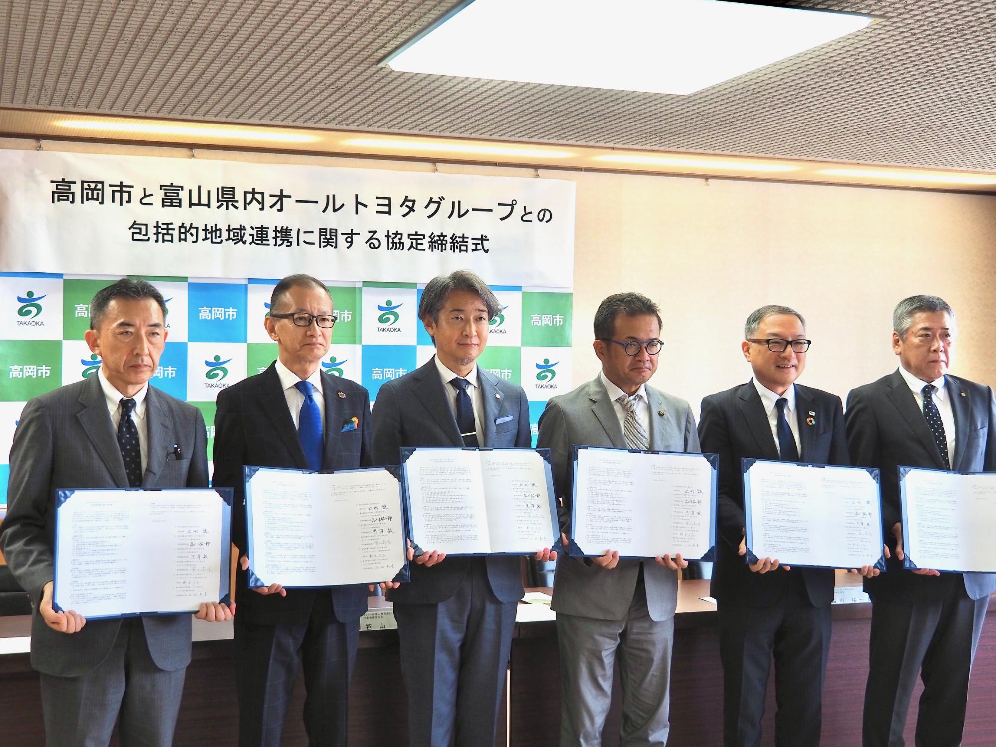 高岡市のロゴが入った衝立の前で、オールトヨタグループと高岡市の代表者が、報道関係者に向かって協定の証書を広げて見せている写真