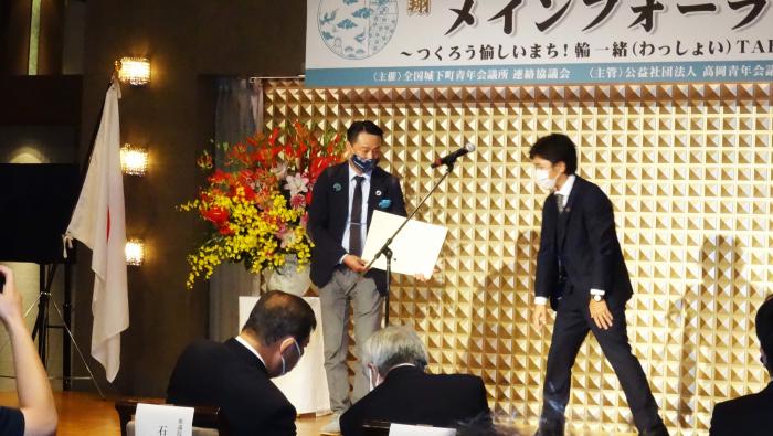 有限会社モメンタムファクトリー・Oriiの折井宏司さんと1人の男性が壇上に立ち、折井宏司さんが賞状を手にしている様子を写した写真
