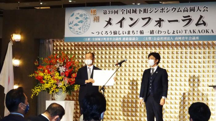 株式会社大越仏壇の大越則夫さんと1人の男性が壇上に立ち、大越則夫さんが賞状を手にしている様子を写した写真