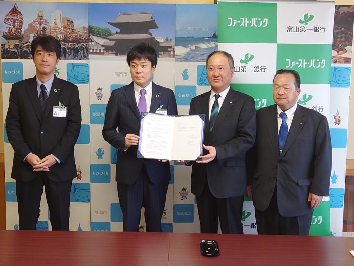 高岡市役所庁議室内で、中央に立っている高岡市長と富山第一銀行の代表者が協定書を2人で持ちながら並び、その両隣に男性2人が立っている様子の写真