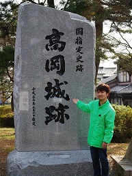 高岡城跡と彫られた大きな石碑を掌で紹介するようにポーズを取る福岡 経さんの写真