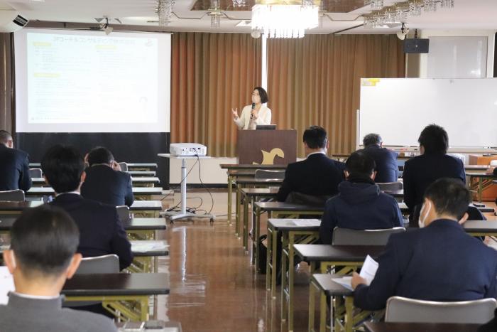 演壇に立った女性が、スライドショーを使用しながら講演をし、参加者が、資料を見ながら熱心に聞き入っている様子の写真