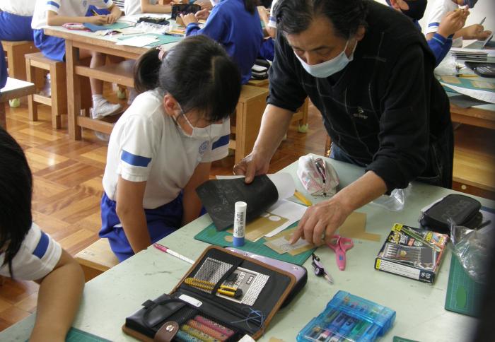 小学校の工作室内にて、職人の指導により、漆器や銅器の作品づくりを行う女子児童の様子を写した写真