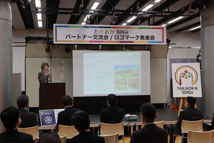 「たかおかSDGsパートナー交流会」と書かれたボードの下で、演台を前にした女性が立って話をし、スーツ姿の男性たちが座ってそれを聴いている写真