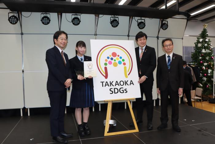 イーゼルの上に置かれた「たかおかSDGsロゴマーク」選出作品のポスターのそばで、スーツ姿の3人の男性と制服姿の1人の少女が微笑んでいる写真