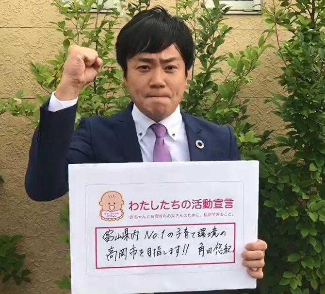 建物を背景に、「わたしたちの活動宣言」と書かれたフリップを左手で持ち、右拳を顔の横に振り上げている高岡市長の写真