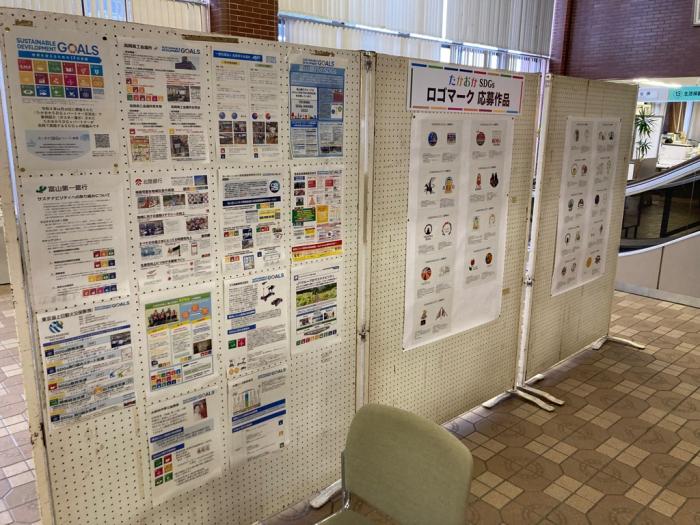 衝立に、「たかおかSDGsロゴマーク」応募作品のコピーと、SDGs活動の取り組みを解説したチラシが、貼られている写真