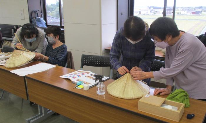並べられた木目のパイプテーブルを使って、菅笠製作を2人一組で協力して行う、高齢の女性達の様子を写した写真