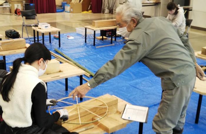床に敷かれたブルーシートの上に並べられた木製の作業台を使って、職人の指導の下、菅笠作りを行う女性の様子を写した写真