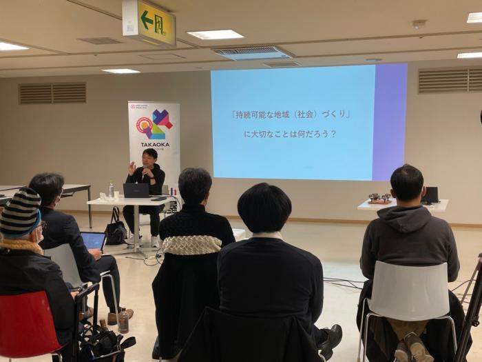 パソコンを開いた机の前に座った男性が、スライドショーを使って、椅子に腰かけた聴講者たちに、講演をしている写真