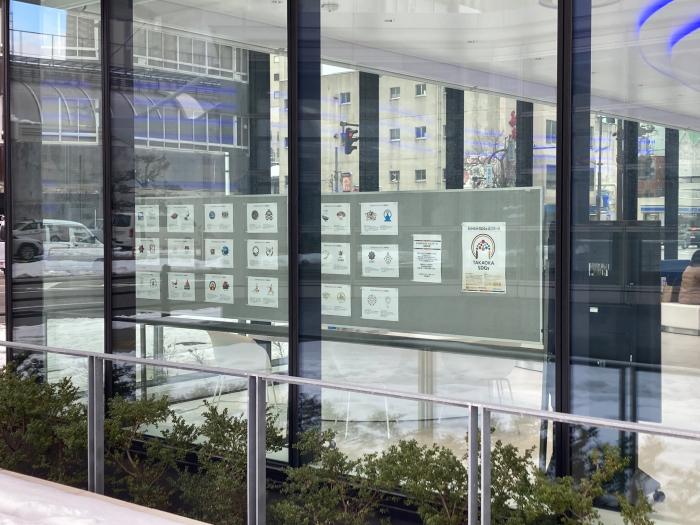 銀行らしい建物の中の衝立に、「たかおかSDGsロゴマーク」選出作品とその他の応募作品のコピーが貼られているのを、外からガラス越しに見た写真