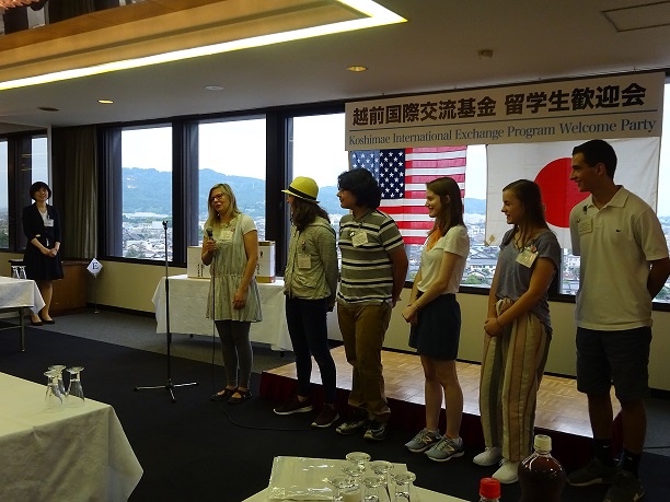 アメリカと日本の国旗が窓ガラスに飾られた前に6人の留学生が立ち、その内の1人がマイクで発表をしている写真