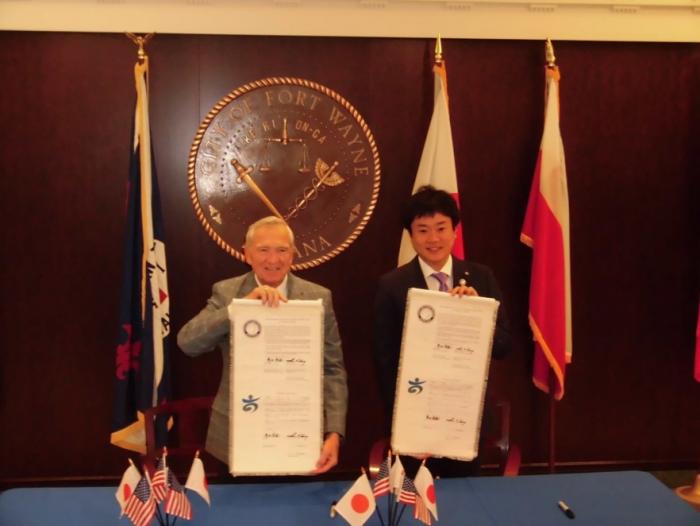 小さな国旗が飾られたテーブルの前でスーツ姿の男性2人がそれぞれ書類を掲げている写真