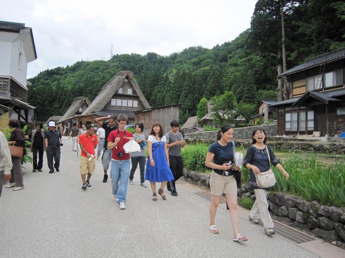 かやぶき屋根の家や木造住宅の側にある道を歩いている人たちの写真