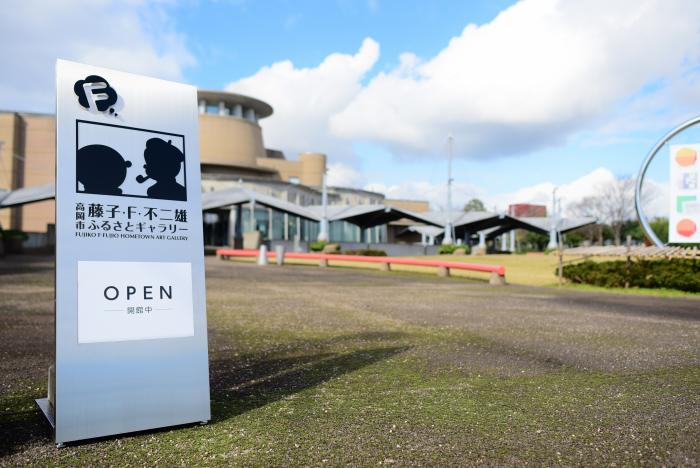 屋外にて高岡市美術館を背景にしつつ設置された、案内用立て看板の写真。看板には市役所前の看板と同じレイアウトのシルエットがプリントれている様が確認できる
