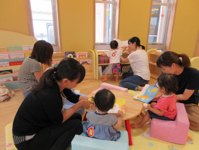 本棚の前に配置された、幼児用の机と椅子に座って絵本を読む子どもたちと、それを横で援助する大人を写した写真