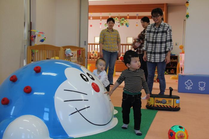 ドラえもんの遊具が前面に置かれ、2人の乳幼児と3人の大人が後ろに立っているところを写した写真