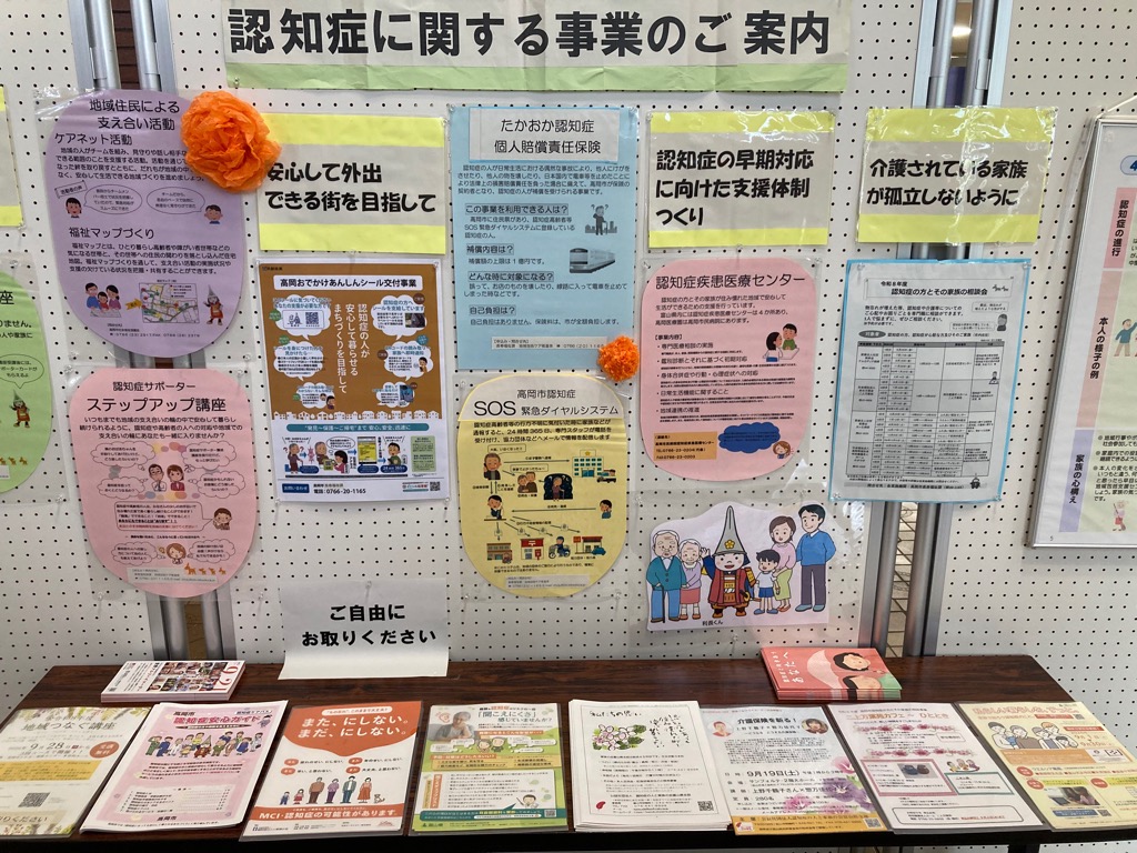 「認知症に関する事業のご案内」と装飾された展示物があり、その手前に関連する資料が並べられている様子の写真