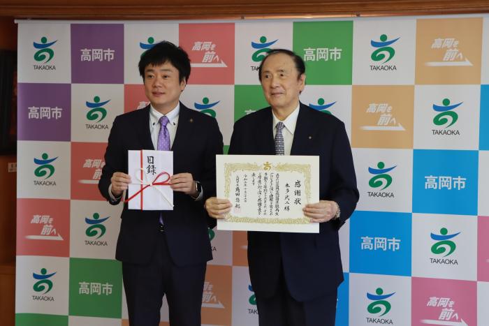 スーツ姿の男性2人がそれぞれ手に目録と感謝状をもって写真を撮られている姿の写真
