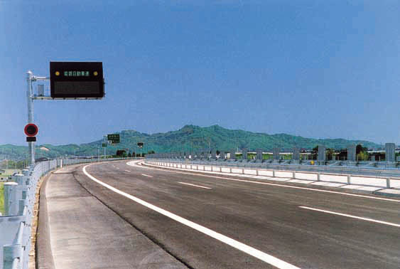 遠景に山を臨む電光掲示板や標識がある道路の写真