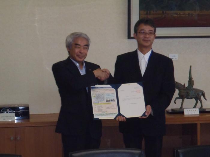 登録証伝達式の様子を収めた写真。高橋市長と入江氏の両名が登録証を掲げつつ、握手を交わして記念撮影に応じる様子が確認できる