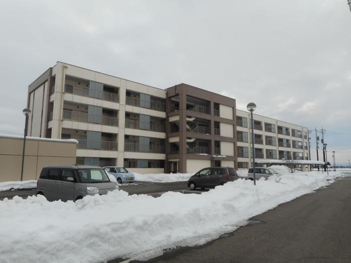 雪の積もった駐車場の奥にある4階建ての建物の写真