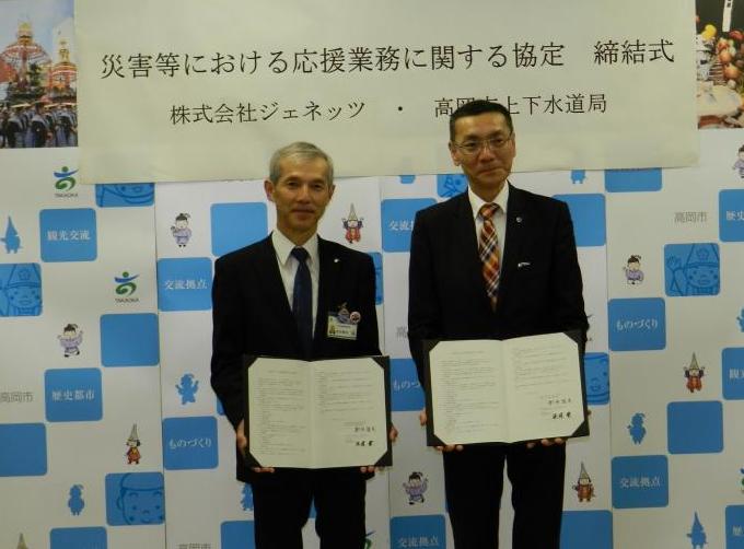 白と青の背景の前に立ち締結式の書類を見せる男性2人の写真
