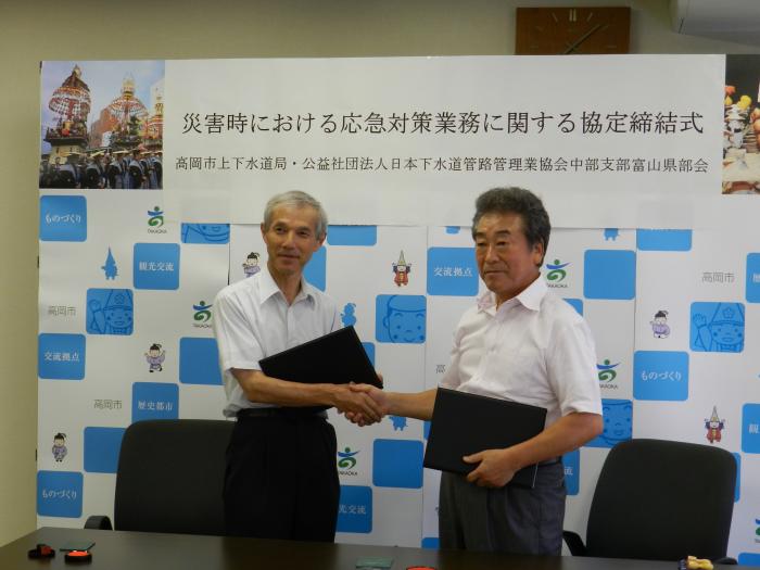 白と青の背景の前で椅子から立ち上がり握手をする男性2人の写真