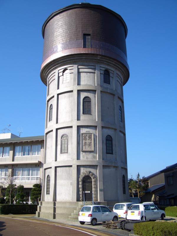 青空の下白い車が3台停まっていてその横に下が灰色上が茶色の塔のような建物の写真