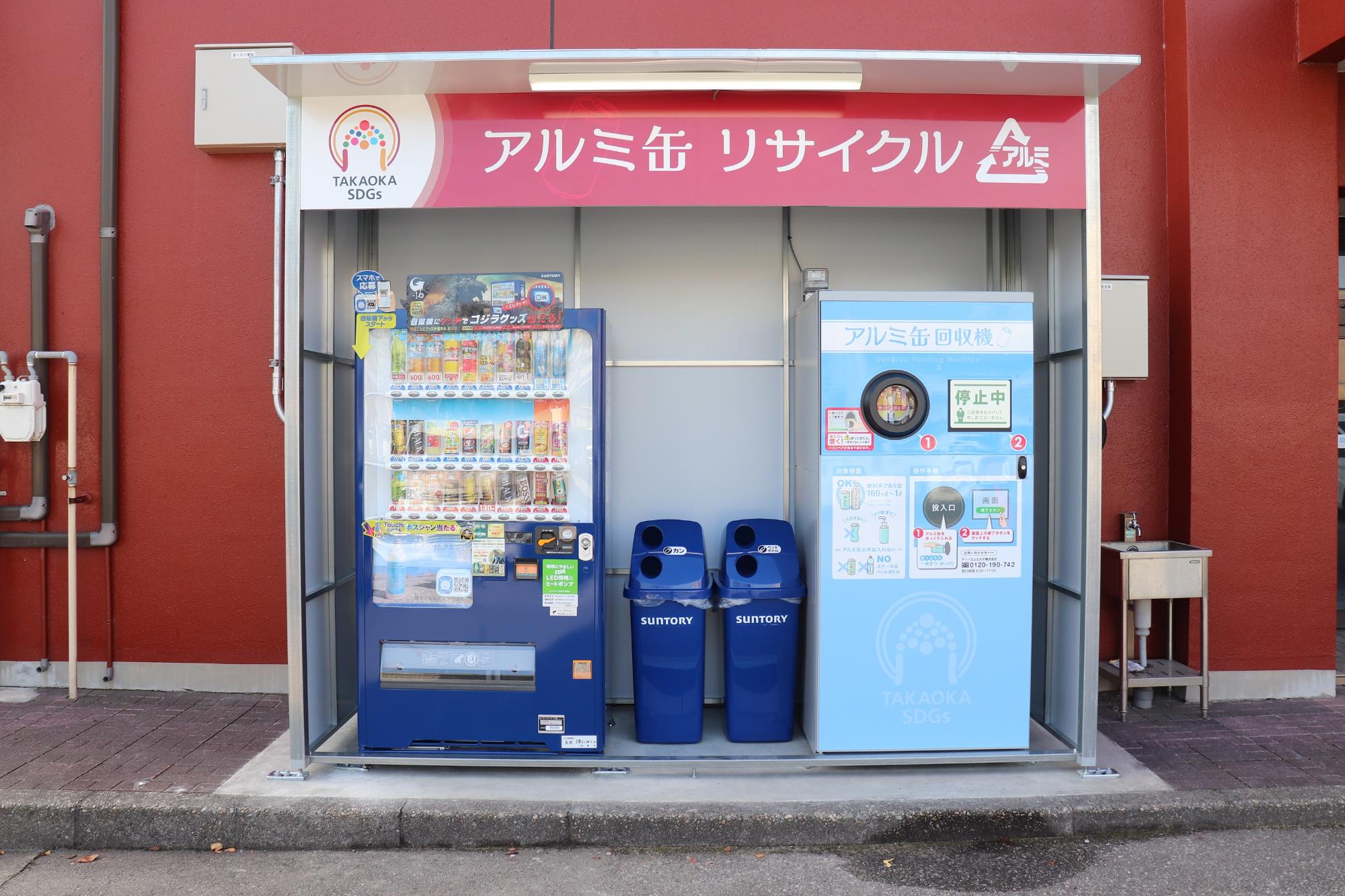 屋根がついた小屋に看板が設置され、アルミ缶リサイクルと書かれており、その下に青色の自動販売機と青色の2つのゴミ箱と水色のアルミ缶回収機と書かれた機械が置いてある写真
