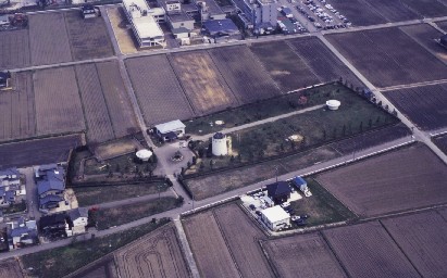 背の低い木に囲まれた野原が田んぼが並ぶ中にあり中央に小さい白い塔がある空から撮られた写真
