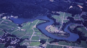 蛇行した形のダムと奥には森が広がり手前には緑の田んぼが並ぶ空から撮られた写真