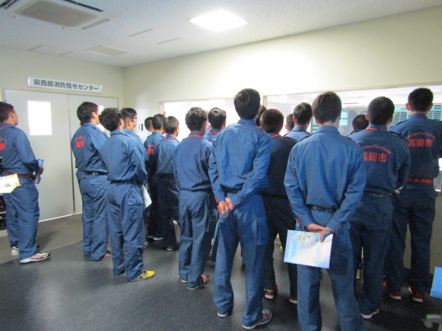 施設内で、作業着を着た大勢の男性がガラス張りの隣の室内の状況を見学しているの様子の写真