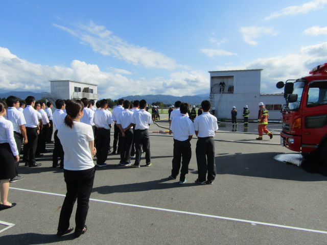野外で、消防車と消防服を着た消防隊員数名が作業している状況を見学する大勢の男女の様子