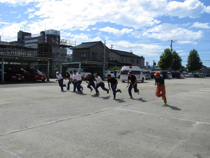 野外の、路面がアスファルトの敷地内にて、9名の帽子をかぶった男性が全力疾走をしている様子の写真