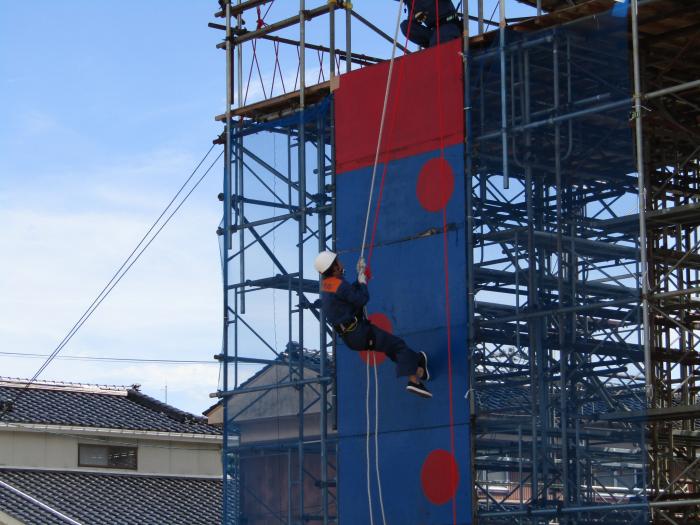 奥に二階建ての民家が見える野外で、その民家よりも高く組まれた鉄骨の足場の上から下に垂らされたロープをつたって登るヘルメットを被った男性の様子の写真