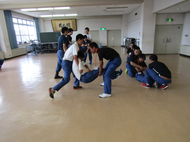 室内で、3人一組になり、1人の男性を2人で道具を使わずに搬送しようとする4組の男性の様子の写真