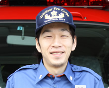 消防車の前に刺繍の入った帽子と襟にバッジのついたシャツをきている小出 拓朗さんが微笑んでいる様子の写真