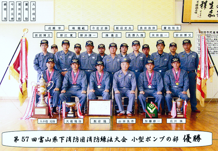第57回富山県下消防団消防操法大会小型ポンプの部優勝と書かれ、優勝旗やトロフィーが並べられた室内で大勢の団員たちが3列に並んでいる様子の写真