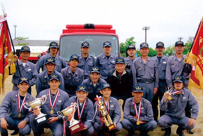 野外で消防車の前に3列に並ぶ18人の消防団員がおり、両脇に立つ団員は優勝旗のような旗を掲げ、前列にしゃがんで並ぶ団員6人はトロフィーやメダルなどを持っている様子の写真