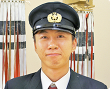 火消し纏を背に、警備服を着た浜谷 大介さんが正面を向きやや笑顔でいる様子の写真