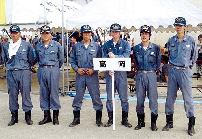 後ろにテントのようなものがあり、その手前に、6人の消防帽子とつなぎのようなものを身に纏った男性のうちの1人が高岡とかかれたプラカードを手に持っている様子の写真