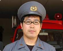 中央にバッジのついた帽子をかぶっている眼鏡をかけた沖田 智則さんが消防車の前に立っている様子の写真